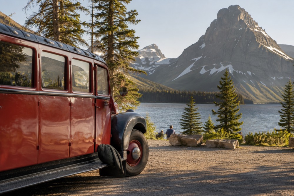 A red vintage tour bus parked beside a lake with mountains behind it