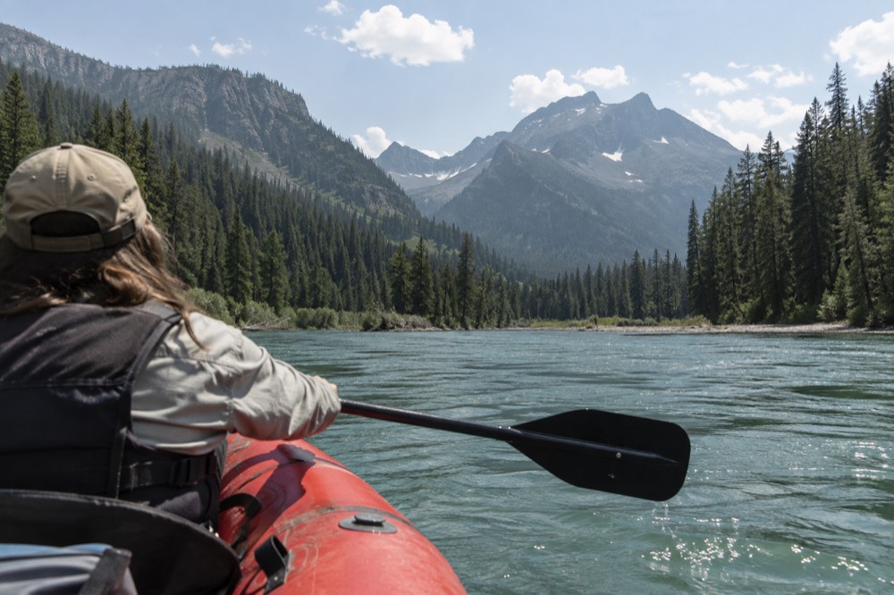 A raft floating down a wide green river between forested mountains on a summer day