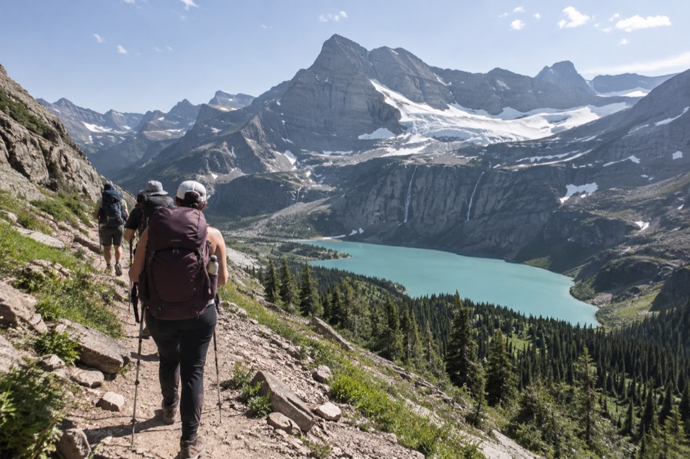 A guided hiking group walking a dirt trail toward a snow-fed lake below tall peaks