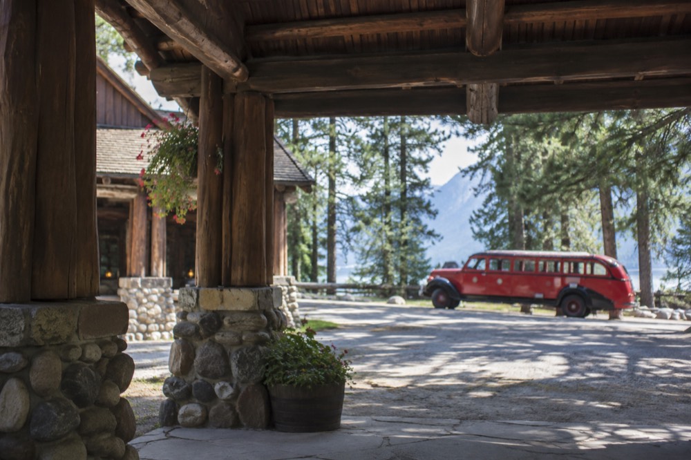 A historic timber lodge with a red bus parked on a mountain road beside a lake