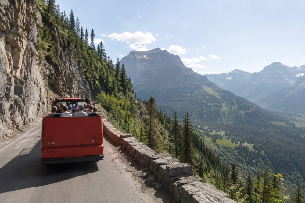 A red open-top tour bus winding along a narrow mountain road with a glacier-carved valley below