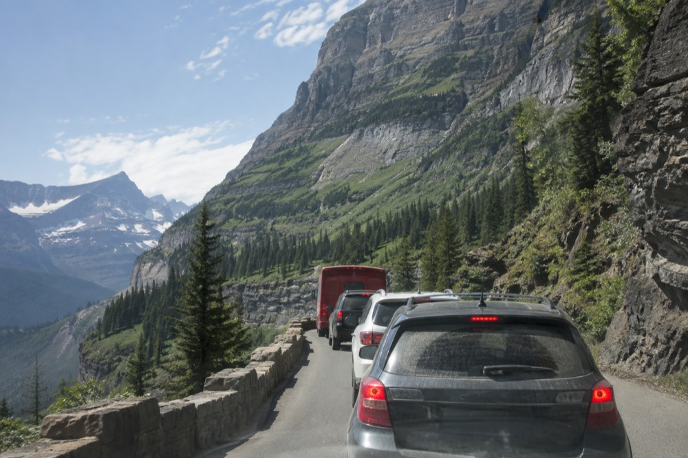 Traffic queued on a scenic mountain road with a red tour bus among the vehicles and cliffs rising above