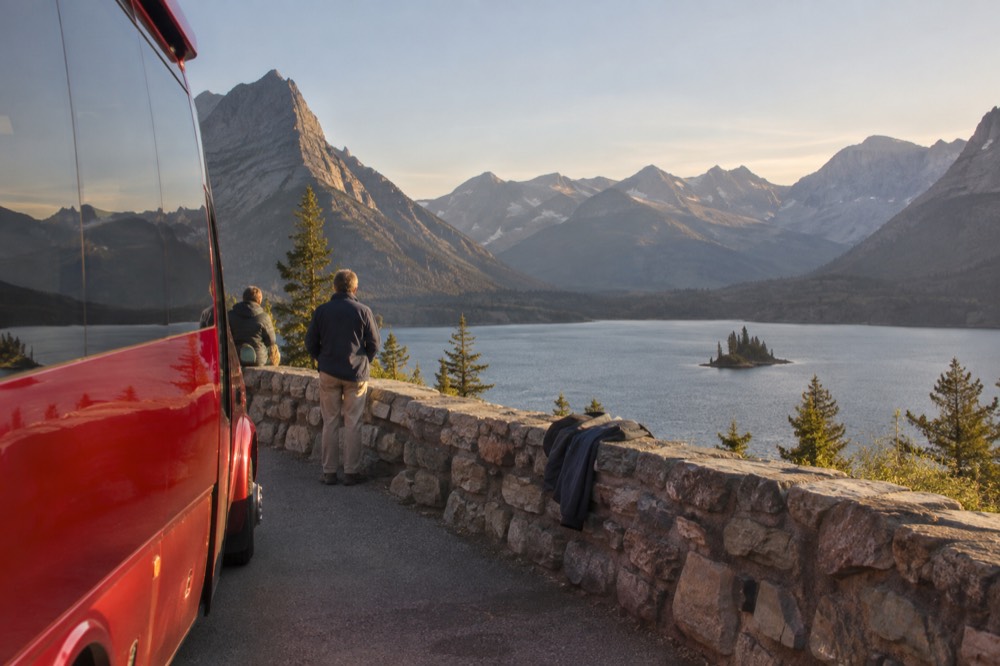 A red tour bus parked at a scenic overlook with passengers' views of a glacial lake and jagged peaks beyond