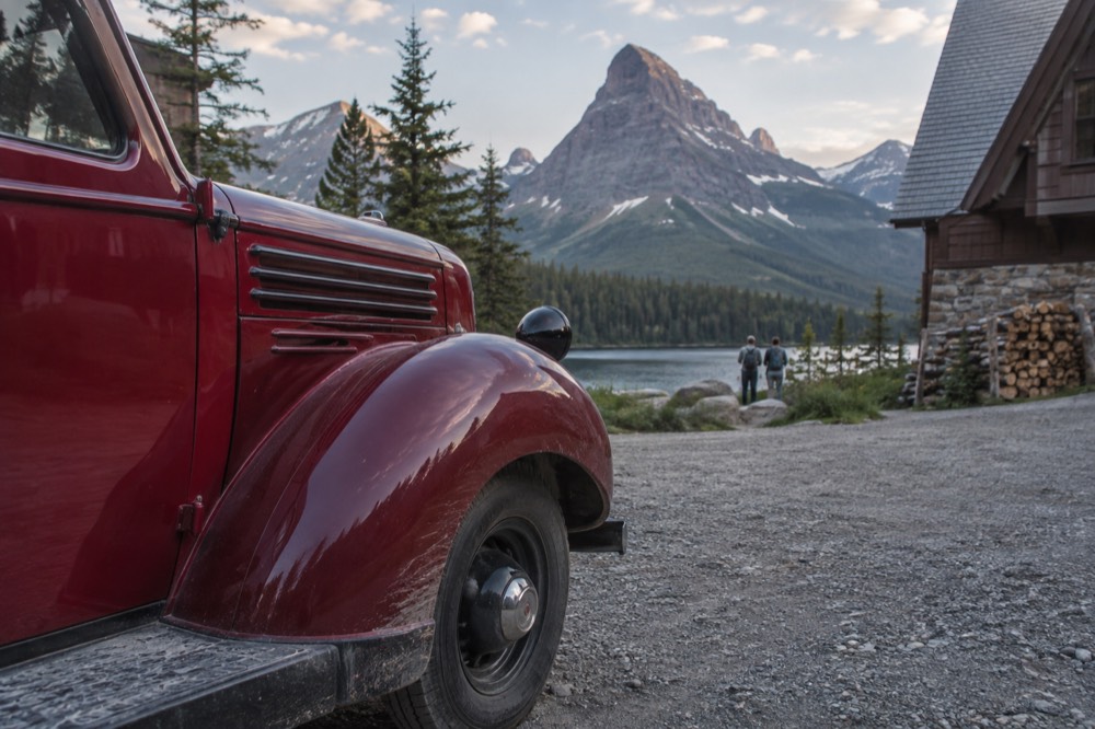 A red vintage tour bus parked beside a calm lake with forested mountains behind it.