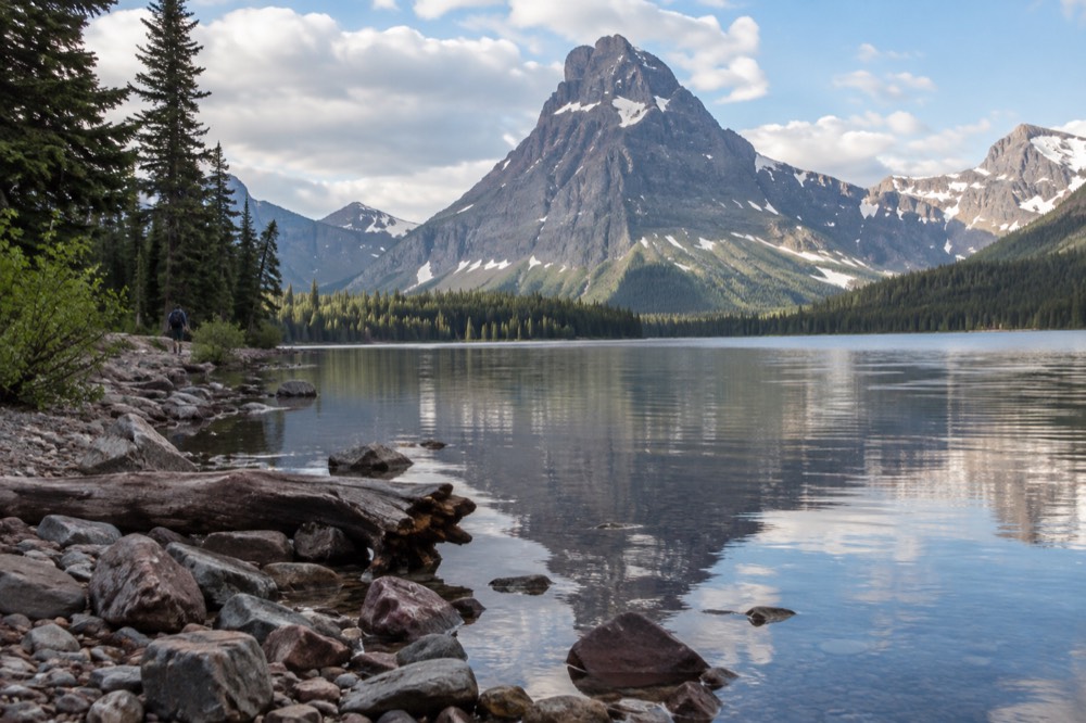 A long alpine lake reflecting jagged snowy peaks under a partly cloudy sky.