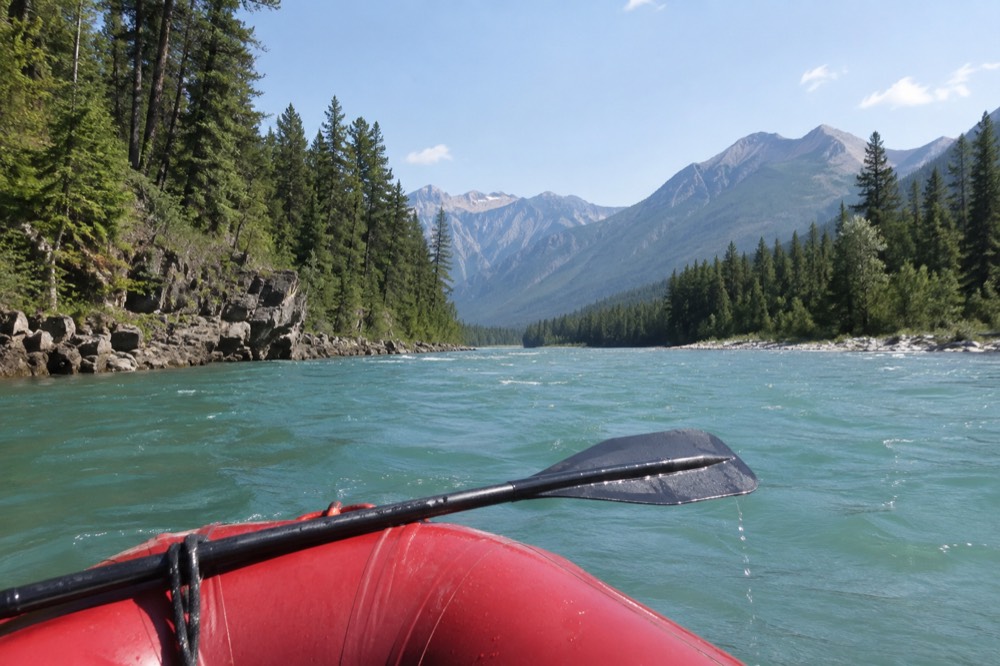 A raft floating down a wide turquoise river between forested mountain slopes
