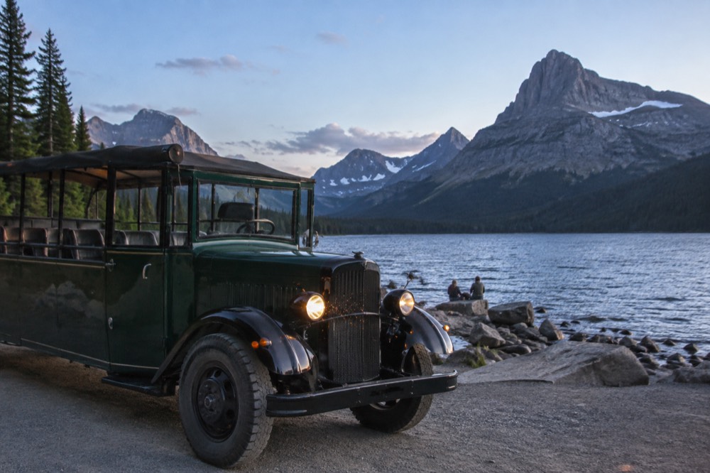 A vintage open top bus parked beside a lake with tall mountains behind it