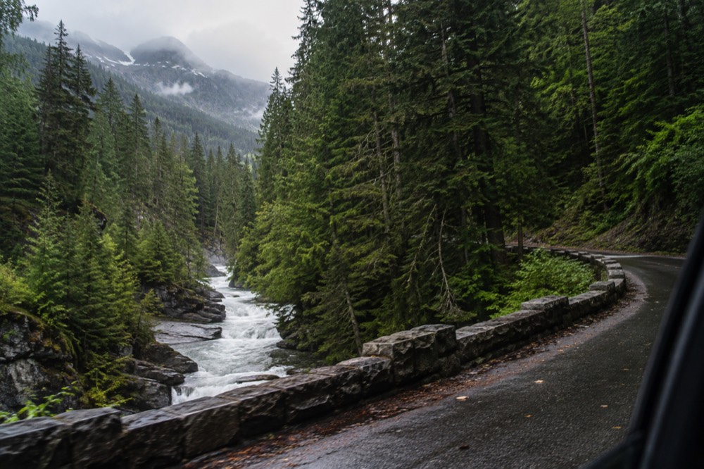 A winding mountain road with tall peaks and dense forest on either side
