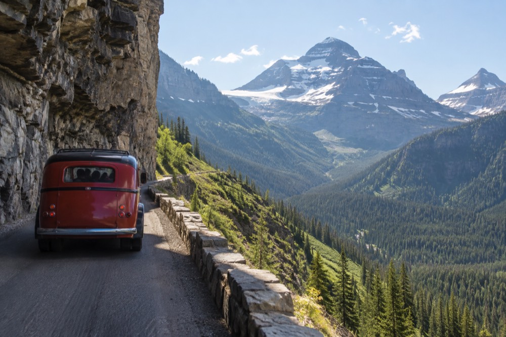 A red vintage tour bus on a narrow mountain road with steep drop-offs and distant peaks