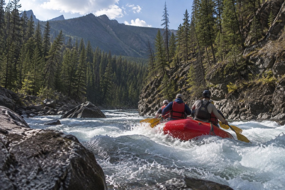 A raft full of people paddling through whitewater rapids with forested mountains behind