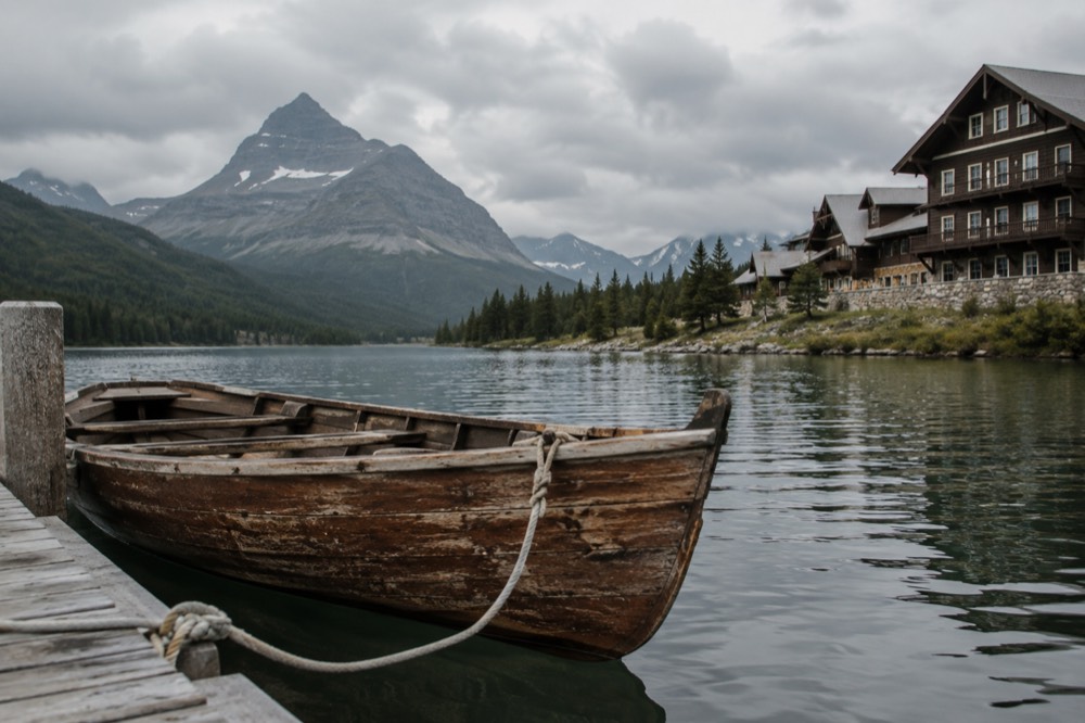 A historic wooden boat docked at a lakeside pier with mountains reflected in still water