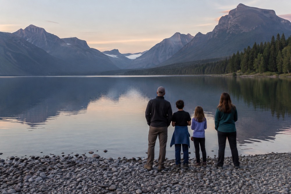 A family standing at a lake shore viewpoint with mountains reflected in still water