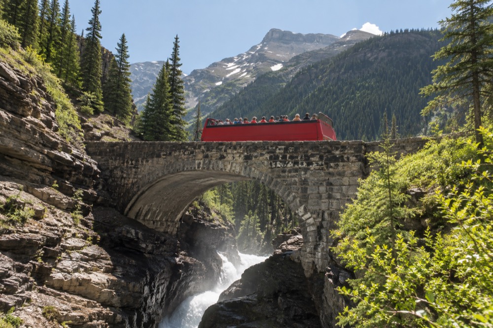 An open-top red bus crossing a stone bridge above a river gorge in mountains