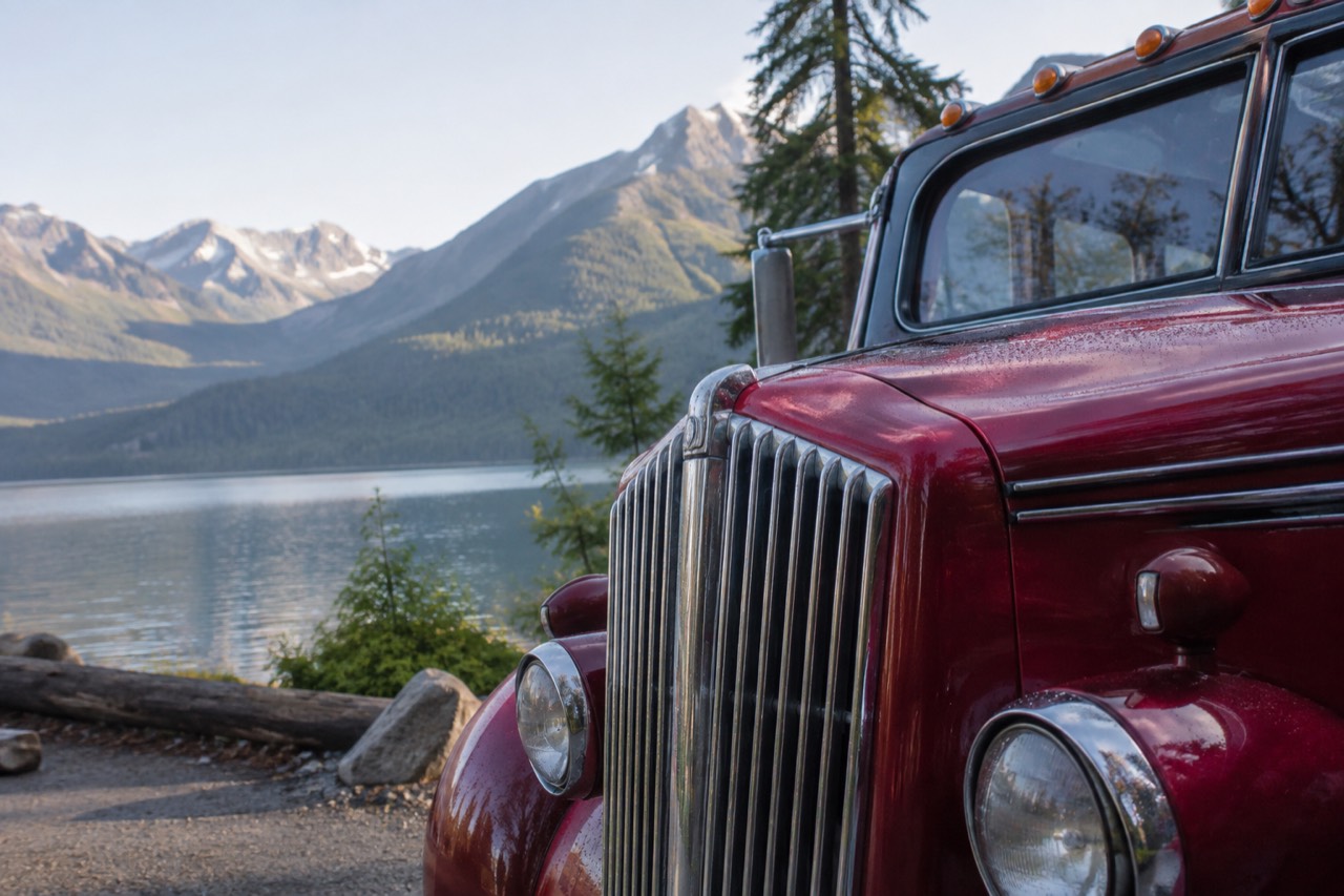 A vintage red touring bus parked on a mountain road beside a lake and forested peaks