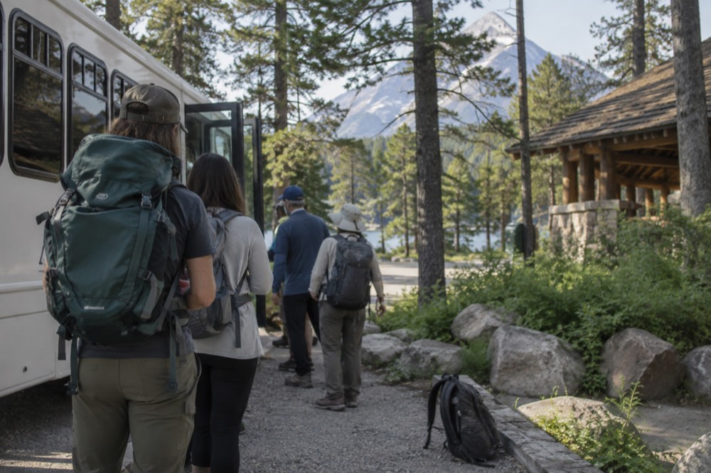 A line of visitors waiting beside a parked shuttle bus at a wooded trailhead