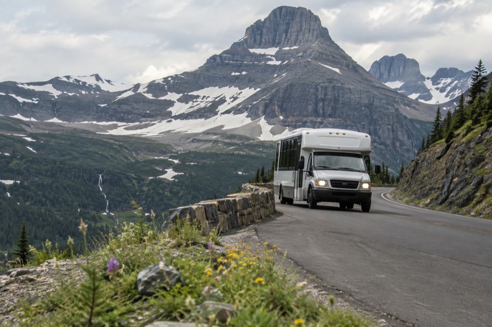 A shuttle bus rounding a curve on a high mountain road with snowfields above