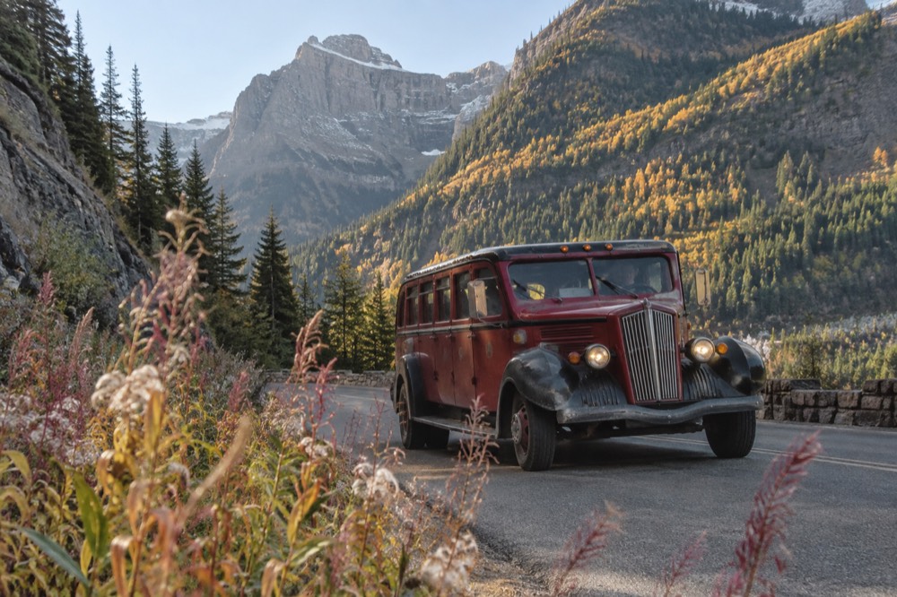 A red vintage tour bus rounding a curve with mountains in the background
