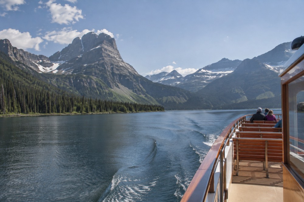 A historic wooden tour boat with an open upper deck approaching a forested shoreline