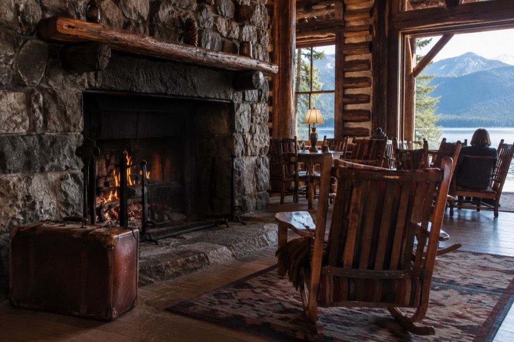 A wooden lodge with a large stone fireplace and rocking chairs facing a lake view