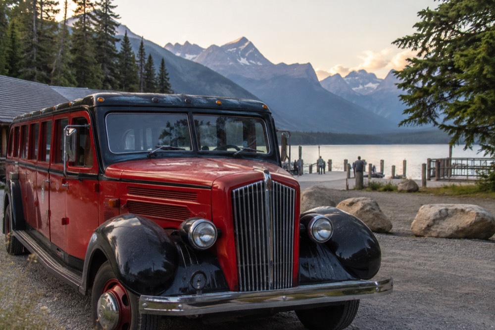 A red vintage tour bus parked beside a mountain lake with forested peaks behind it