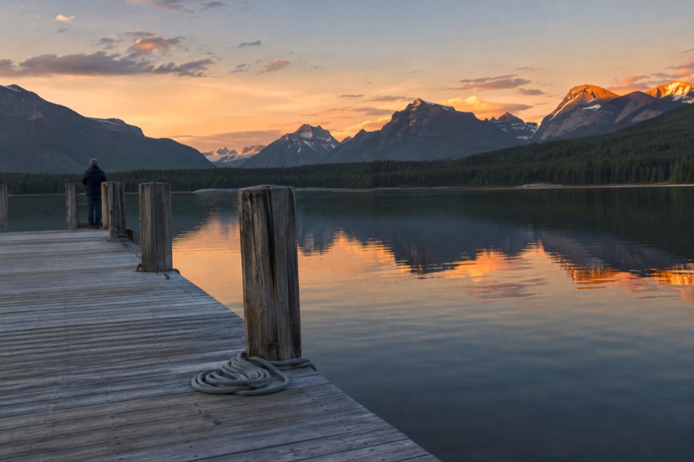 A calm lake reflecting mountains at sunset with a wooden dock in the foreground