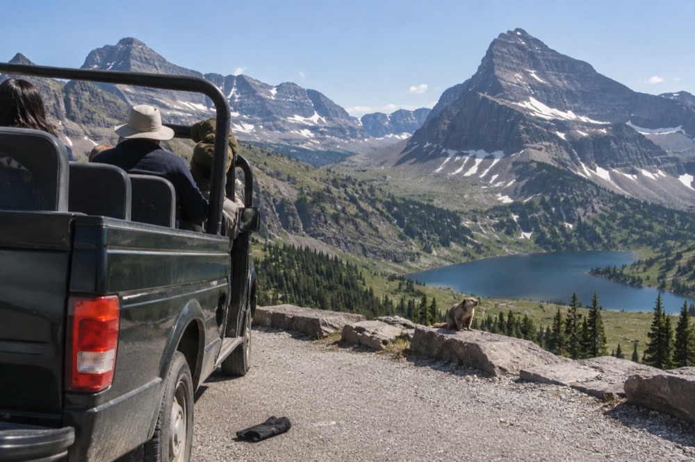 An open touring vehicle parked beside an alpine lake with peaks behind it