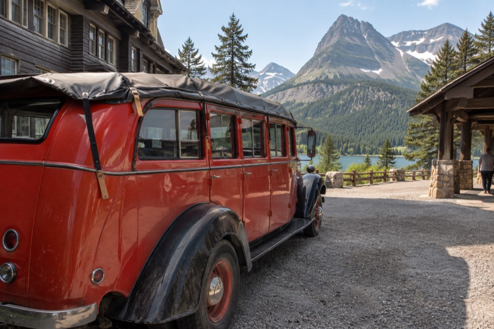 A red historic touring bus parked beside a lake with forested mountains behind