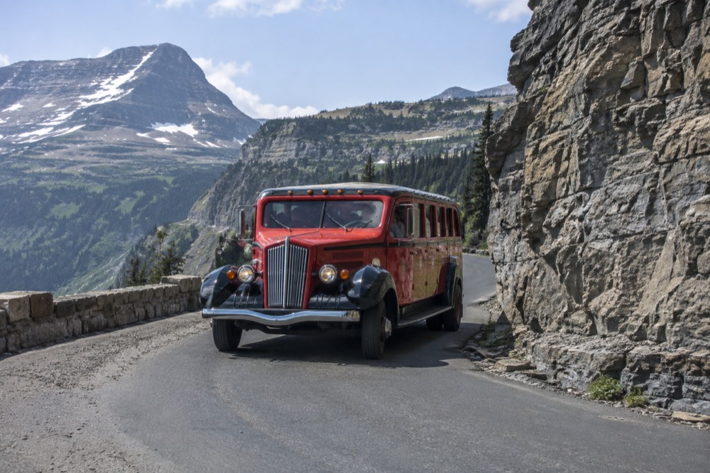 A red vintage touring bus rounding a curve on a mountain road with a tall rock wall on one side