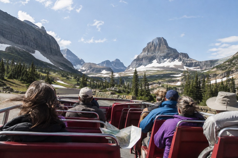 A red open-top touring bus parked at a high mountain pass overlook with snowfields and jagged peaks beyond