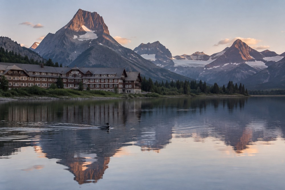 A mountain lake with a historic wooden hotel on its shore and steep glaciated peaks reflected in still water