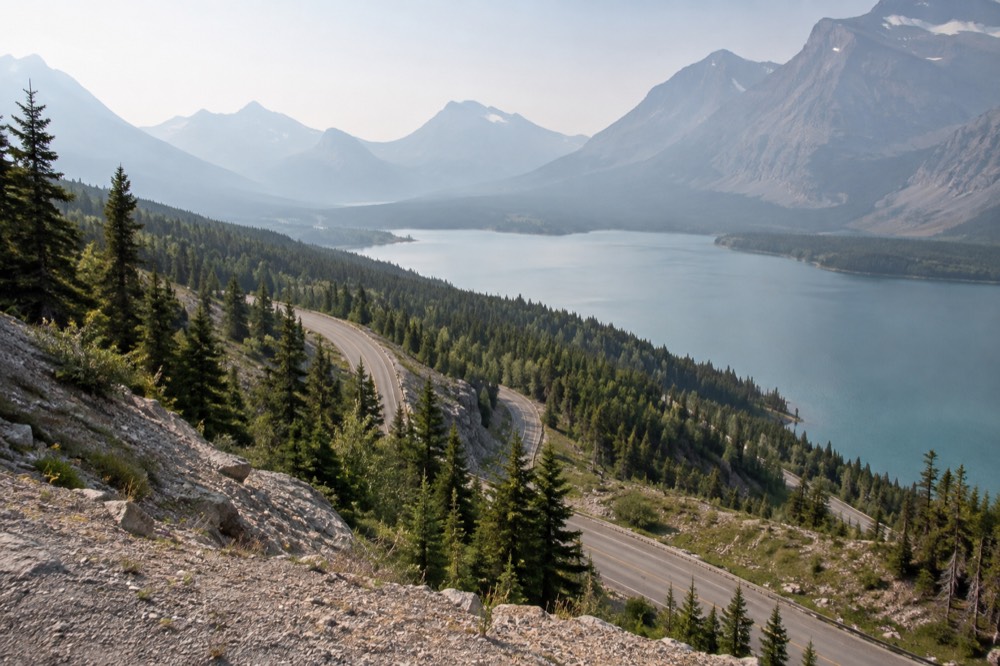 A winding mountain road descending toward a turquoise lake with forested slopes on either side
