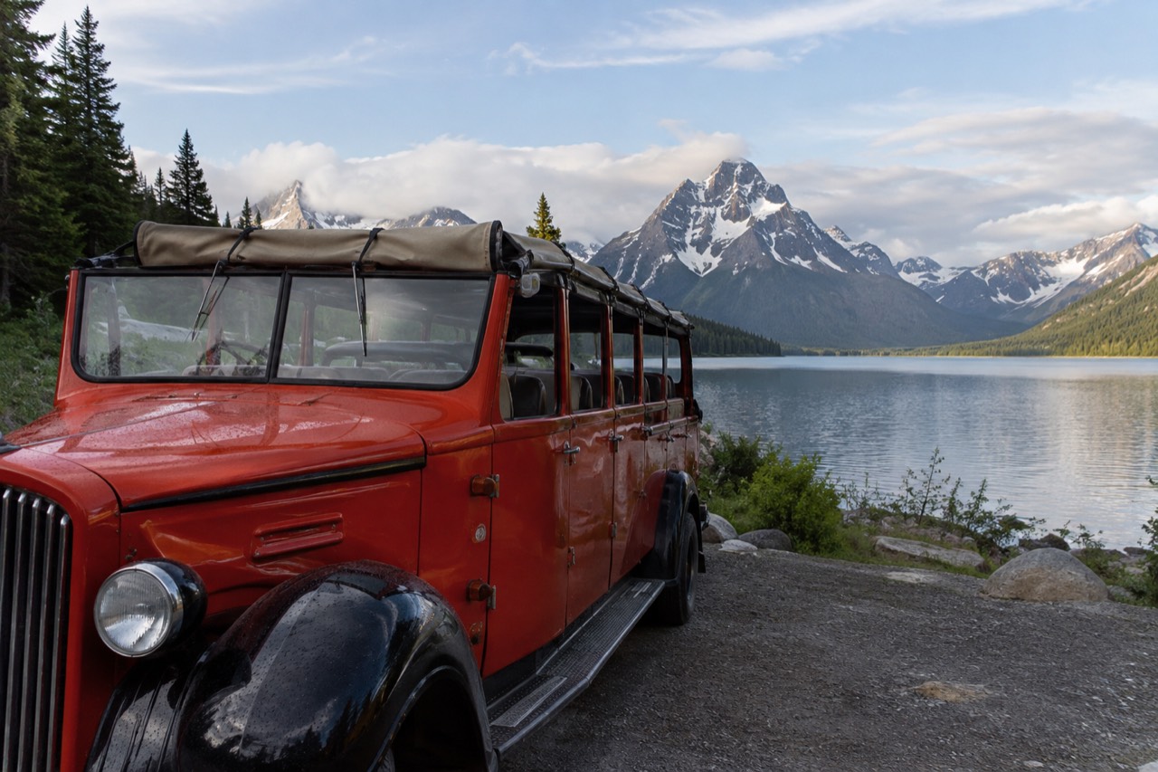 A vintage red touring bus with its canvas top rolled back, parked beside a mountain lake with snow-capped peaks behind it