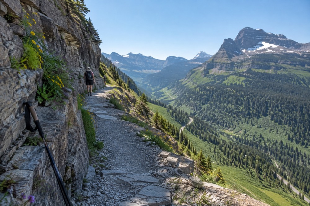 A cliff-hugging trail along a green mountainside with a road visible far below
