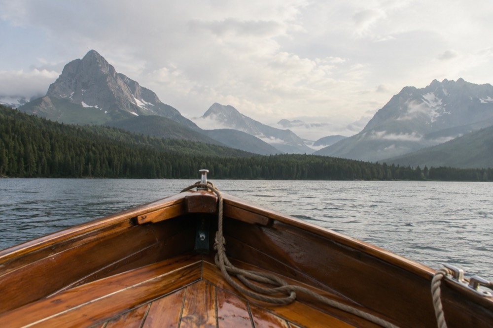A wooden historic boat crossing a calm lake toward jagged peaks