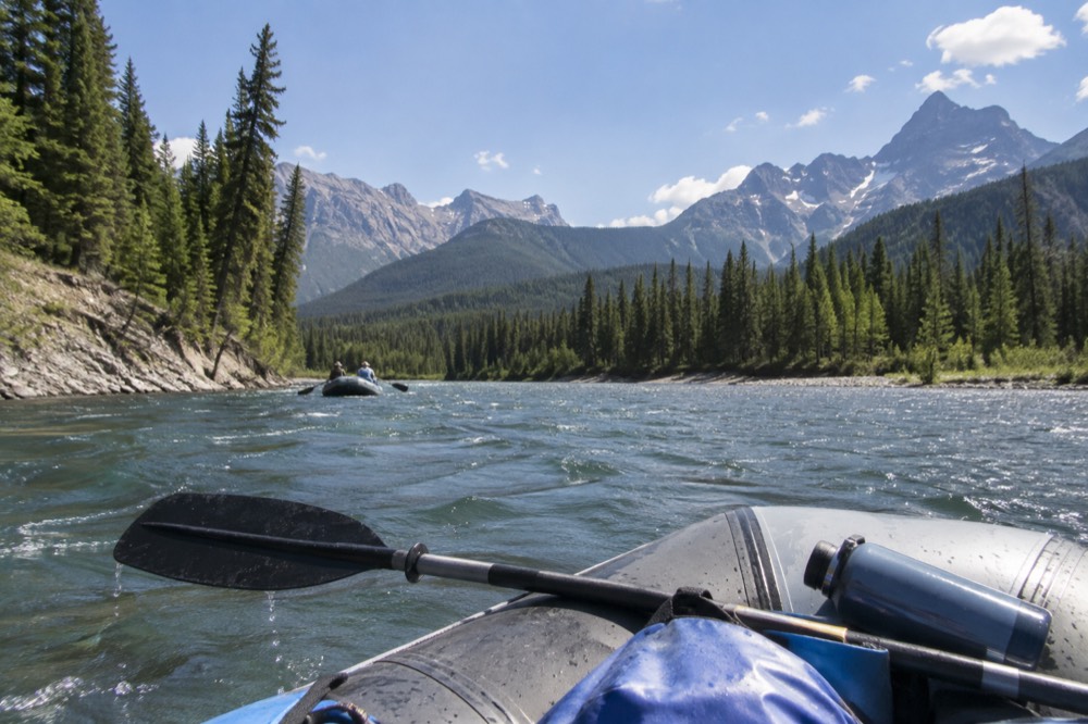A raft floating down a wide river between forested banks under summer sky