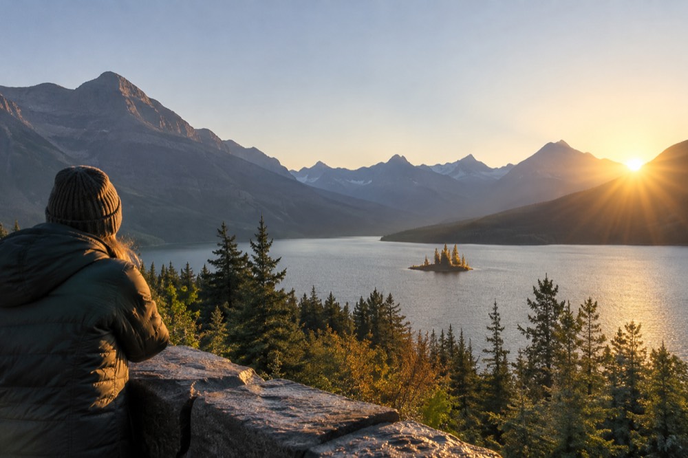 A small forested island in a turquoise lake beneath tall mountains at sunrise