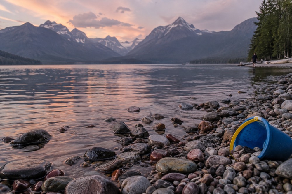 A lakeshore beach with smooth pebbles, calm water and mountains in the distance