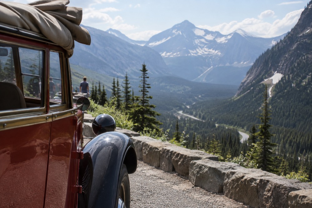 A vintage open-top tour bus parked beside a mountain road overlooking a glacial valley