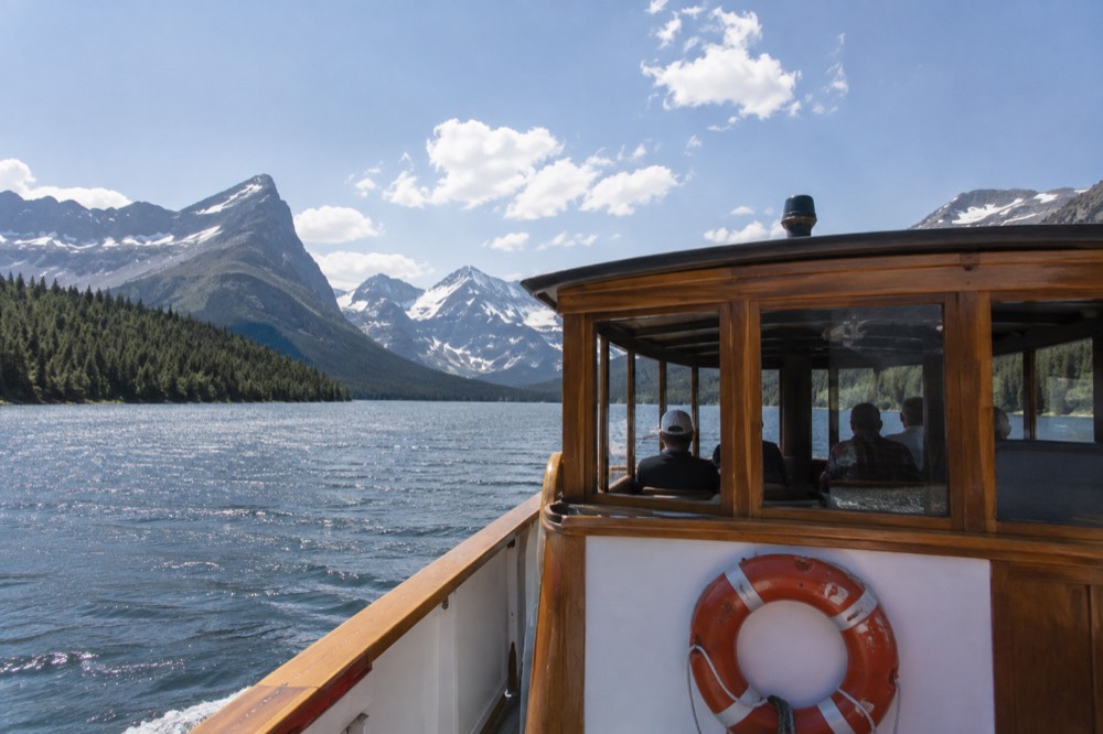A wooden boat cruising across a wide turquoise lake toward jagged snow-capped peaks