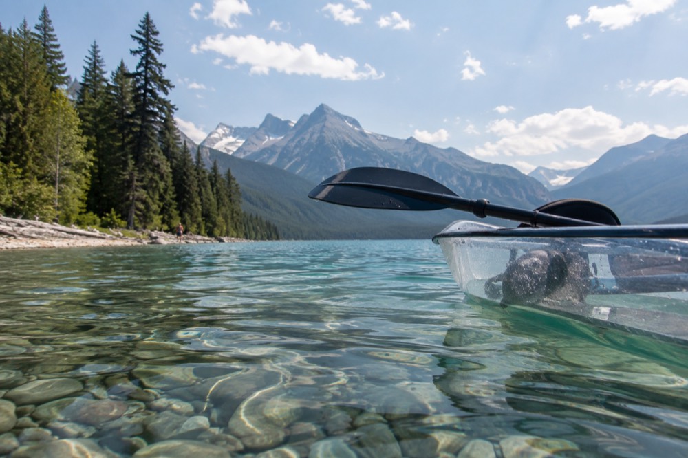 A clear kayak gliding over shallow turquoise water with a forested shoreline in the distance