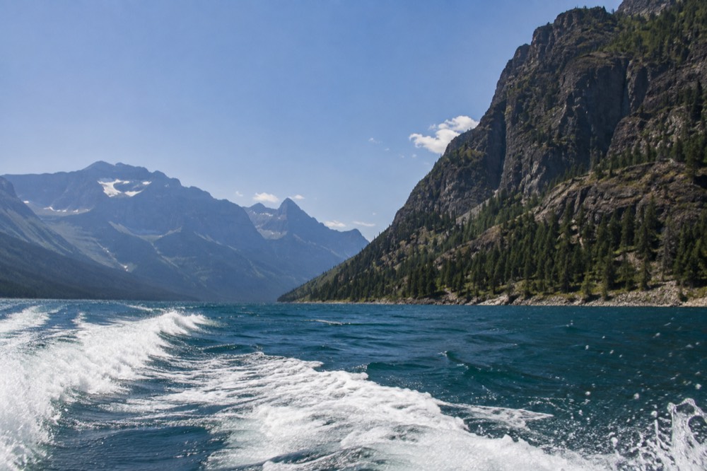 A motorboat wake crossing a deep blue lake with tall cliffs along the far shore
