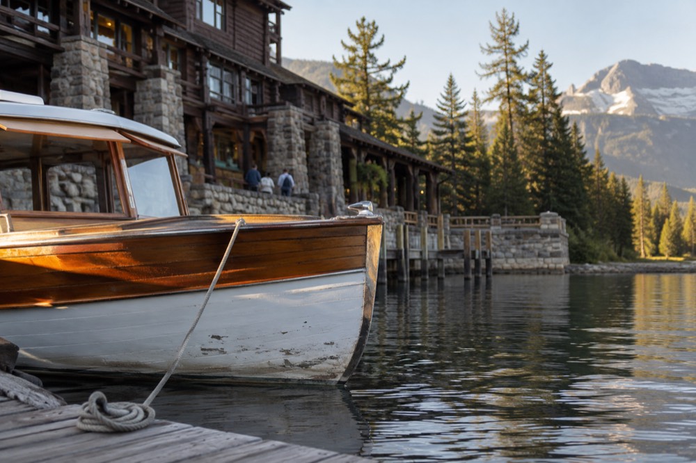 A historic wooden boat docked at a stone lodge with a lake and mountains behind