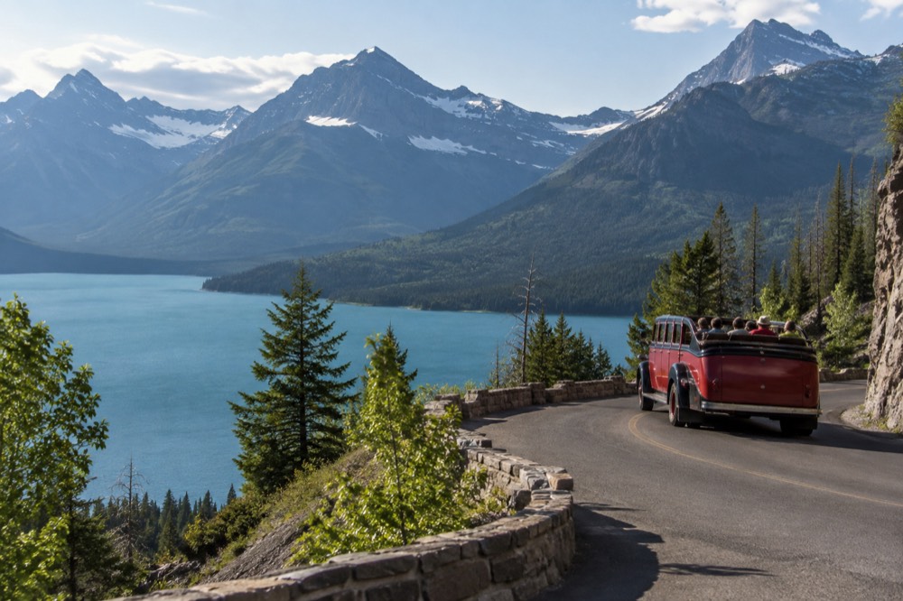 A red open-top tour bus on a curved mountain road with a glacial lake in the valley below