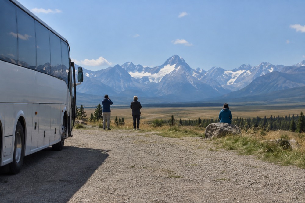 A coach bus parked at a scenic overlook with wide prairie meeting a wall of mountains