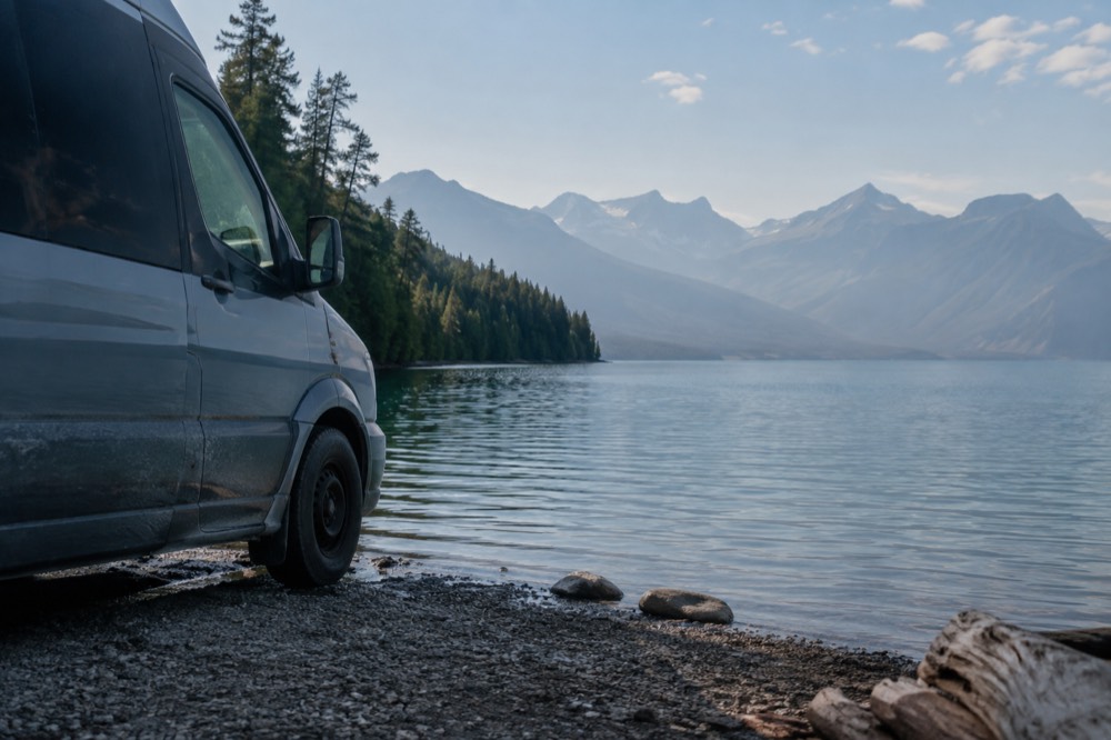 A small passenger van parked at a lakeside turnout with a long blue lake and mountains beyond