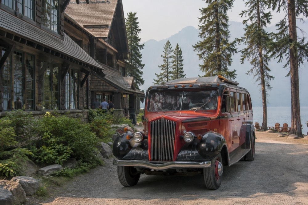 A red vintage tour bus parked beside a mountain lake with forested slopes behind it