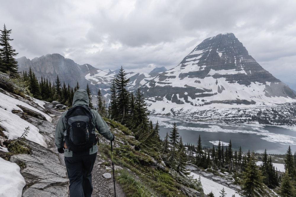 A hiker on a narrow trail with a mountain vista and patches of snow