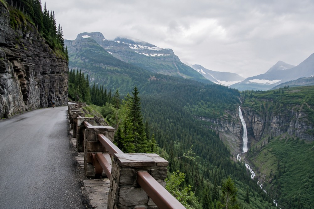 A narrow mountain road curving along a steep rock wall with a waterfall across the valley