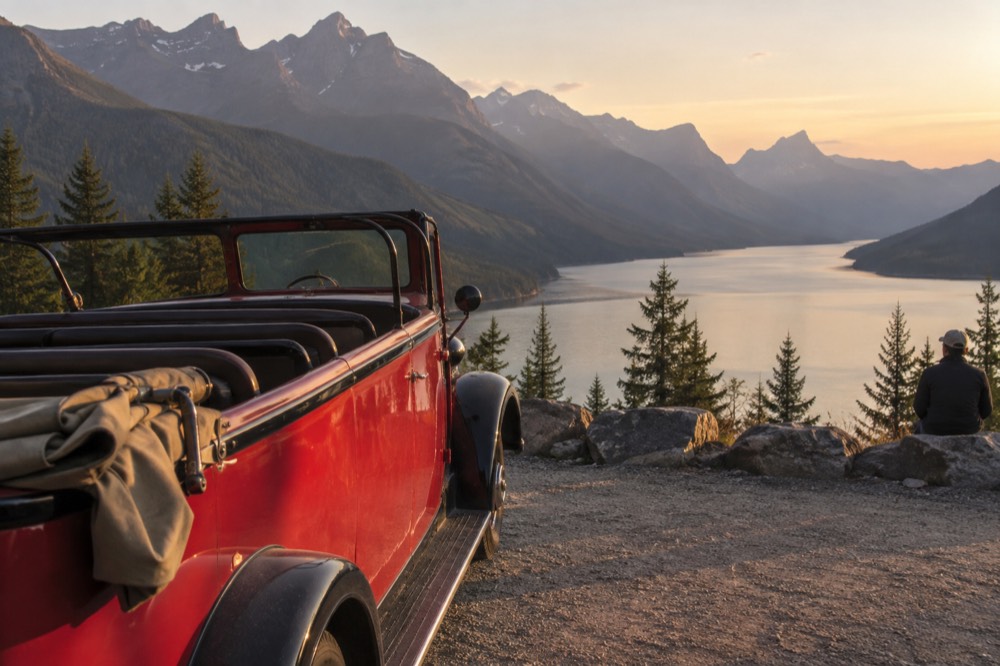 A vintage open-top tour vehicle parked at a mountain overlook with a long lake below