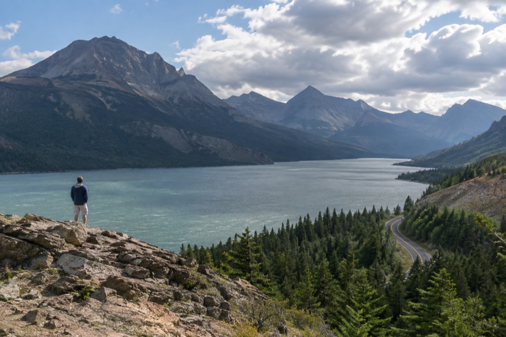 A winding mountain road above a turquoise lake with forested ridges in the distance